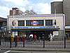 A grey-bricked building with a rectangular, dark blue sign reading "MILE END STATION" in white letters all under a light blue sky with white clouds