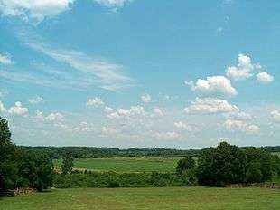 Photo shows a rural scene under partly cloudy blue skies. In the foreground a grassy hillside slopes down to a tree-filled ravine. On the other side are more open fields, with trees in the distance.