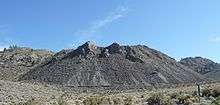 Steep-sided large pile of dark gray sharp rock in a field of scrub vegetation.