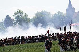 Photo shows a re-enactment of a Napoleonic battle. A French firing line is seen from the rear while a church tower looms to the right.