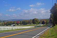 A rolling landscape with hills in the distance, a line of trees some of which are turning red, and fields in the foreground. A white fence separates them from a road that stats at bottom left and then passes between trees at the right center before cresting. At the bottom right is a small green sign with white numbers on a metal stand.