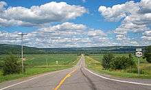 A two-lane paved road stretching straight ahead, going slightly downhill to a level section across fields towards a ridge in the rear. At the right of the image is a sign with "EAST 258" on it.