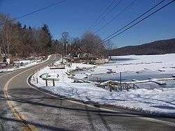 A road straddling the bank of a frozen lake. The surrounding ground is covered by thin layer of snow. Powerlines are visible overhead