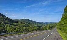 A two-lane road descending the right side of a wooded valley under a blue sky with minimal clouds