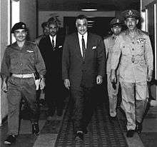 Three important men walking in a hall, the first and the third are in military garb, the second is in a suit and tie. Behind them are three other men