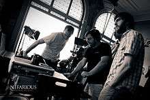 A black-and-white photograph of four men standing in a room filled with equipment including a videocamera, a speaker, and a spotlight