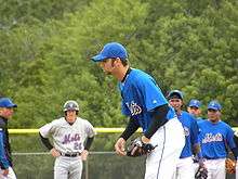 A man in a blue baseball jersey and baseball cap and white baseball pants bending over from the waist; he wears a black baseball glove on his left hand