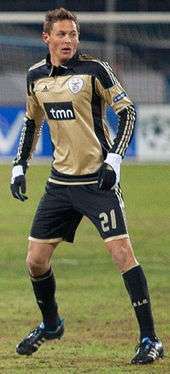 A young man on a football pitch during a game. He is wearing a bronze jersey with black details, and black shorts, socks and gloves