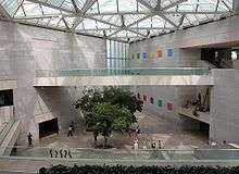 A large open cement room contains several people on the ground far below a balcony. Several trees are planted in the concrete floor, and an array of clear windows let in sunshine from above.