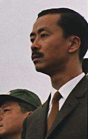  A middle-aged man with side-parted black hair and a moustache, in a black suit, white shirt and brown tie. To the left is a clean-shaven Asian man with black hair and a green military cap.