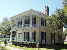 Two storied square-shaped, flat-roofed house with large windows framed by shutters; both second story balcony and first floor porch have Grecian columns.