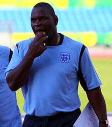 Heavily built middle-aged black man wearing a football shirt with a badge showing three lions