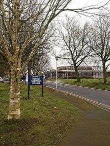 Building with multiple glass windows seen at the end of a tree lined road. Sign says welcome to Norton Radstock College