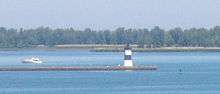 A pier with a square, black and white-striped lighthouse at the end. A small motorboat is paralleling the pier, heading away from the lighthouse.