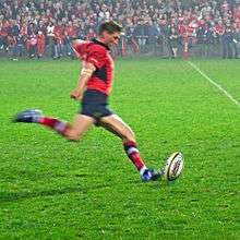 A man in a red shirt is running forward to kick a rugby ball placed on a tee.