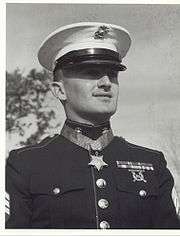 Head and shoulders of a white man with a pointed mustache, wearing a star-shaped medal on a blue ribbon around his neck.