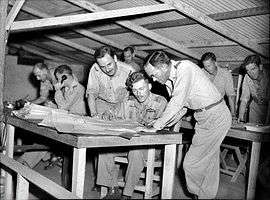 Approximately eight men in military uniforms leaning over papers on desks