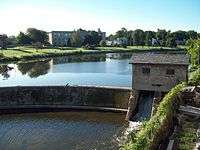 A body of water with a curved stone dam. At the right of the dam is a small stone house, underneath which water flows. There are buildings on land behind the dammed water