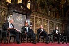 Four men in suits sit on chairs on a red stage in front of an ornate gold and brown wall.