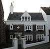 Front view of a dark stone house slightly set back from a street corner. A tiled roof with a dormer window and chimney sits above a white-painted parapet, and six irregularly spaced windows also have white surrounds. The wide, pale blue door, offset to the left, has pillars on each side and a white diamond panel above.