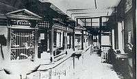 A black-and-white photograph of a bank lobby, with barred teller's windows at a counter on the left and windows and other furniture on the right. A partition is visible in the rear.