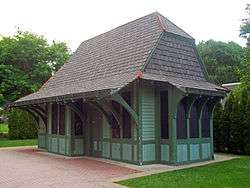 A blue and green wooden building with a curving, pointed roof and tall, narrow boarded-up windows with a brick surface in front and shrubbery behind it