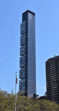 A tall, thin building with some slight squarish projections at its higher levels seen from between some trees.