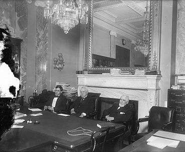 Three white men in suits sitting at a table, with a chandelier hanging and a mirror in the background