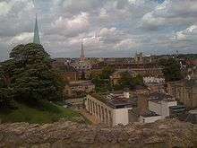 A scene of rooftops; on the left, a grassed mound with a tree; a rough stone wall in the foreground