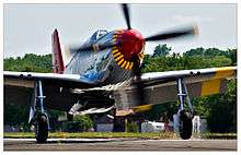 A single nosed-propeller aircraft is on the ground on its wheels with the propeller in motion.  The aircraft is viewed from the front, but the red nose faces slightly to the right.  The aircraft has black propellers.  Parts of the wings and propellers are cropped from view.