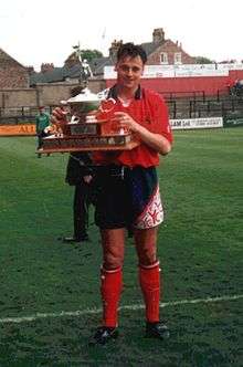 A man with dark hair who is wearing a red top, navy blue shorts and red shorts. He is standing on a grass field, holding aloft a trophy.