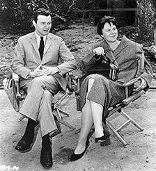 A black and white photograph of Alan J. Pakula seated next to Harper Lee in director's chairs watching the filming of To Kill a Mockingbird