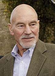 A head shot of a bald hair man with a brown blazer and light blue shirt.