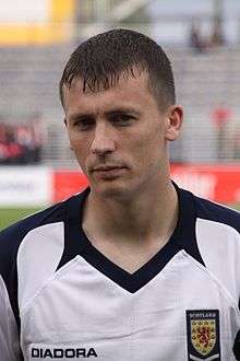 Head and shoulders of a young dark-haired white man wearing a football shirt with Scotland badge