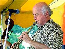 A man wearing a patterned dress shirt, performing a wind instrument while standing behind a microphone