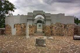 Frontal view through two partially preserved gates of a stone building, with a well in the middle of a courtyard in front