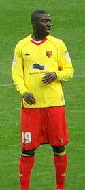 A young man wearing a yellow shirt, red shorts and red socks, standing on a grass field