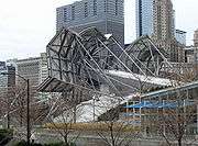 View of the back of a building where many large metal struts support angular metal plates, braced with geometric grids. There are skyscrapers in the background.