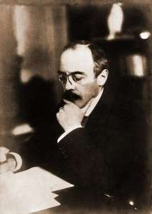 A middle-aged man wearing a suit and spectacles is seated at a desk, examining some papers.