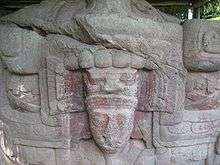 A carved boulder, showing a human face surrounded by elaborate sculpted decoration