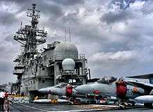 Two pale gray jet aircraft parked on deck of aircraft carrier, with the ship's island in the midground towards left