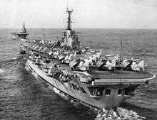 Photograph of an aircraft carrier from behind. Numerous aircraft with their wings folded are sitting on the flight deck. A second aircraft carrier is in the background, leading the first.