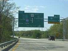 Ground-level view of two lanes of a divided freeway; two large, green exit signs are visible, and trees surround the freeway on both sides.