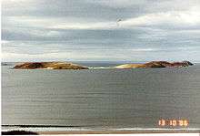 Two green sun-lit islets connected by a sandy tombolo sit offshore from a beach. The skies are grey and the sea all but flat calm.