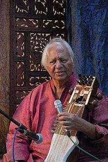 An old man sitting in front of an ornamented partition wall speaks into a microphone and holds a bowed instrument.