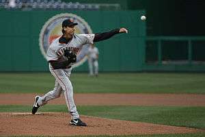 A man wearing a black baseball cap with an interlocking "SF" and grey baseball jersey with "SAN FRANCISCO" on the chest releases a pitch from his left hand.