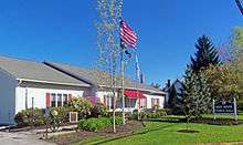 A one-story white building sided in clapboard with red shutters. The American flag flies from a pole in front, where a sign says "Red Hook Town Hall."