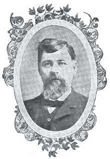 Head of a white man with bushy beard and thick hair wearing a suit, vest, and bow tie. The portrait is surrounded by a decorative oval frame.