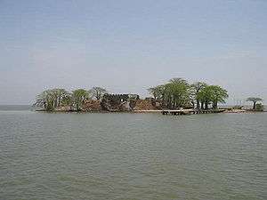 A distant view of very small island home to several thin trees, a brown dock, and a partially obstructed brown shack.