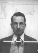 Mug shot of a man with glasses in suit and tie. The number O-15 appears in front of him.